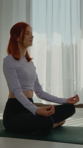 Vertical Screen: A redhead woman sits cross-legged on a yoga mat in her living room, eyes closed, practicing meditation and deep breathing to achieve mental balance during the morning light.