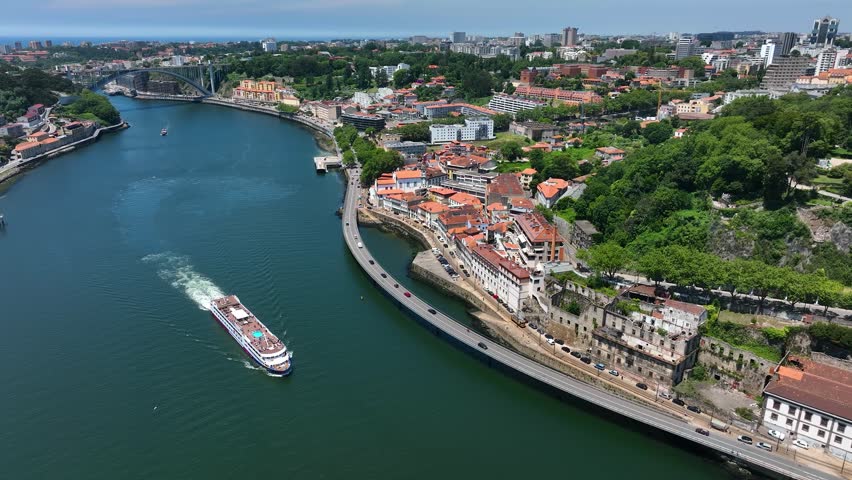 Aerial video of a river cruise ship navigating the Douro River near the riverside buildings and green hills of Porto, Portugal