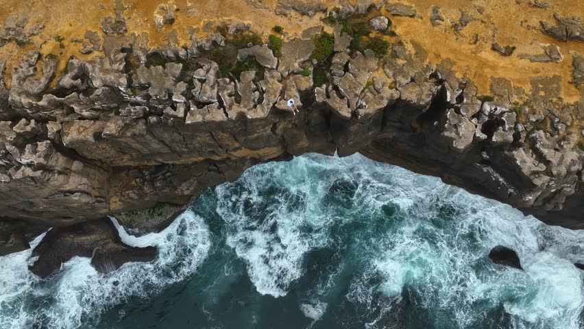 Aerial video of a man sitting at the edge of a rugged coastal cliff in Portugal, overlooking the dramatic waves crashing below against the rocks