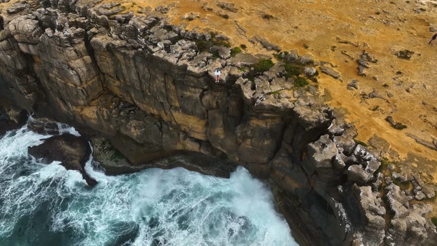 Aerial video of a man sitting at the edge of a rugged coastal cliff in Portugal, overlooking the dramatic waves crashing below against the rocks