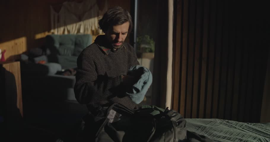 Brunette guy puts his things in a black hiking backpack while sitting on the bed and getting ready to go out in the morning in a country house