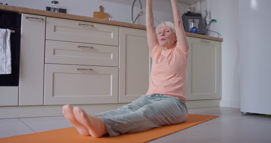 Elderly woman with white hair sitting on orange exercise mat doing stretching and reaching for her legs during her exercise and stretching routine in cozy kitchen at home