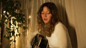 Warm lights illuminate a young woman with long, curly hair, dressed in a simple white shirt, as she plays an acoustic guitar and sings with closed eyes in a cozy, intimate setting - Powered by Shutterstock - Get 15% off with code: PIKWIZARD15