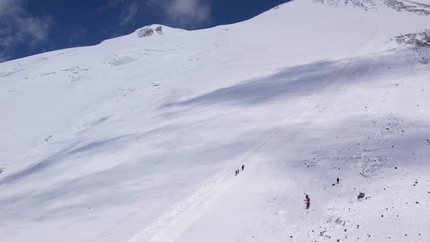Mountaineers ascending a snow-covered path towards the peak of Mount Elbrus. The icy slopes glisten under a deep blue sky as they make their steady climb.