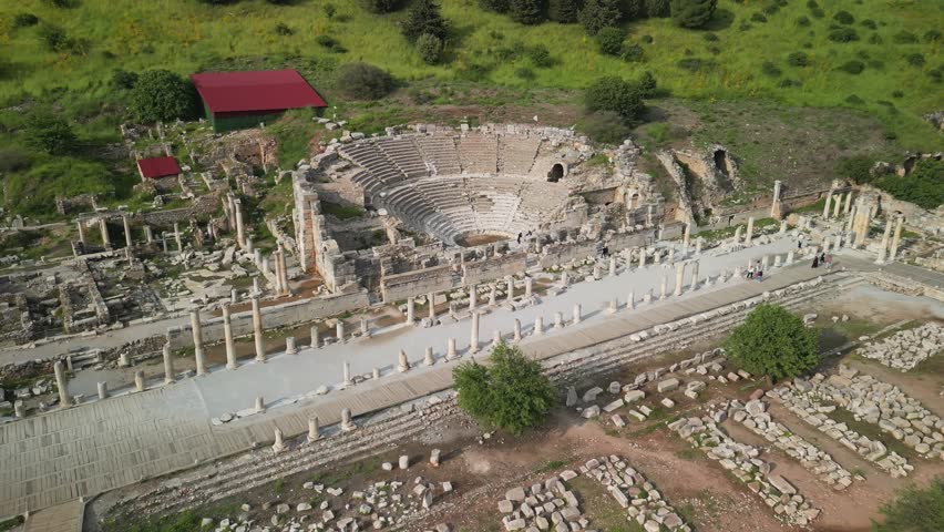 Descending aerial view toward Ephesus small theater in Turkey. Highlights architectural details, surrounding ruins, and green hillside backdrop. A moment captured in historical depth.