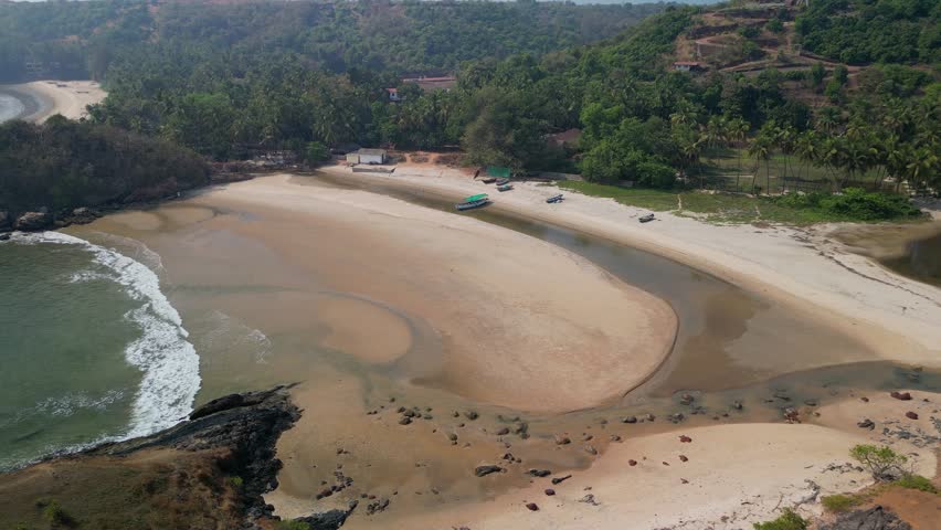 Beaches curve beside forest in Goa. A shallow river joins the sea. Boats rest on dry sand. Palm trees cluster behind. A blend of land and water in a quiet tropical paradise forms