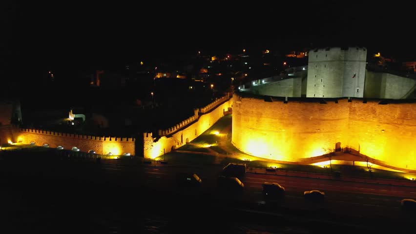 Side perspective of a large fortress in Turkey. Thick defensive walls lit in warm tones. Flag seen flying. Modern road nearby shows contrast between past and present elements.
