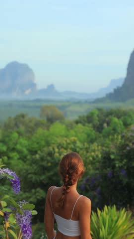 Caucasian woman is seen from behind standing quietly among tropical greenery and purple flowers, admiring Krabi s iconic limestone mountains. Vertical format ideal for mobile ads or social content.