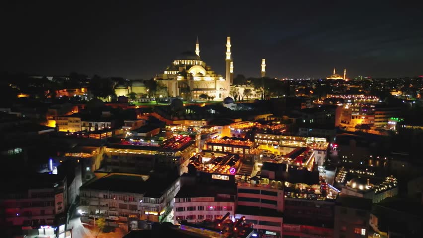Drone flies toward and over Suleymaniye Mosque in Istanbul. Night lights accentuate its majestic Ottoman design, surrounded by vibrant urban life.