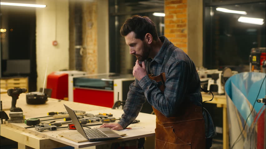 Cheerful Caucasian man wood master carpenter using laptop in workshop builder searching information in Internet guy male chatting social media network pc repairman smile workman order materials online