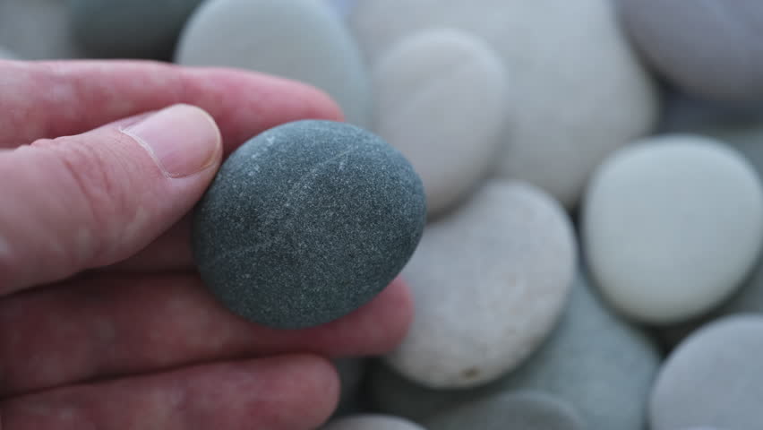 A man holding a pebble in his hand over a background of sea stones. Close up.