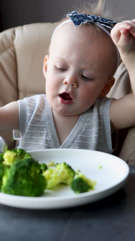 Adorable little baby girl practicing self-feeding with spoon, sitting in high chair, learning to eat healthy broccoli by herself at table