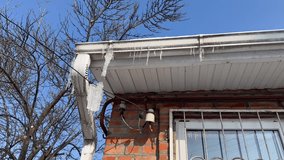 Icicles hanging from the roof of a brick house with visible wiring and window bars. Clear blue sky and bare tree branches indicate cold winter day. Relevant for winter safety and seasonal changes - Powered by Shutterstock - Get 15% off with code: PIKWIZARD15