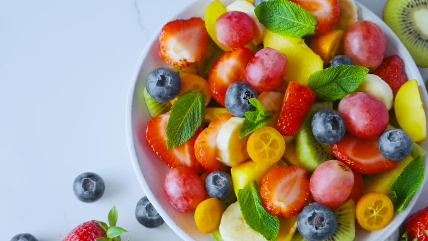 Hand takes strawberry with fork from bowl of summer colorful fruit salad. Top view. Healthy food for breakfast. Mixed strawberries, grapes, banana, kiwi, blueberries, peach, citrus