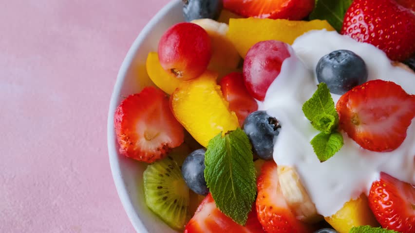 Close up of summer colorful fruit salad with creamy yogurt in a bowl on pink background. Top view. Healthy vegan food with vitamin. Mixed strawberries, grapes, banana, kiwi, blueberries, peach, mint