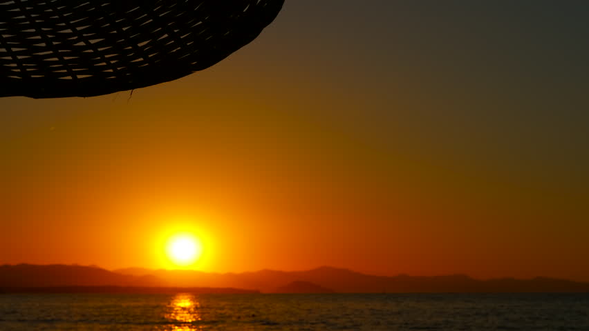 Captivating sunset illuminating ocean waters with warm orange hues. Stunning sunset casts an orange glow over the ocean water as the sun dips below the horizon. A wicker parasol provides shade