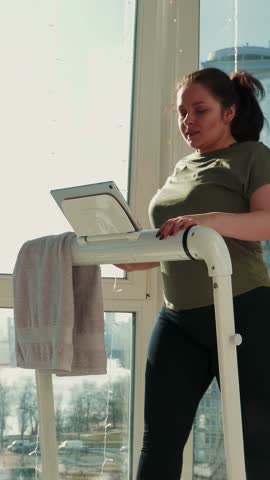 Young athletic woman wipes sweat with towel during hard cardio exercises on treadmill at home. Athletic motivated female is walking on running machine staying healthy at home. Vertical video