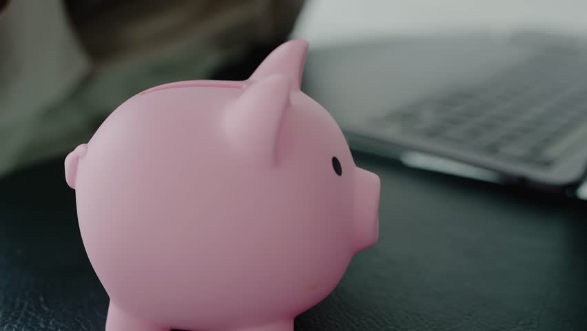 A pink piggy bank with a coin dropping into it near a laptop, symbolizing savings, finance, and money management. Hand Inserting Coin into Pink Piggy Bank with Laptop in Background