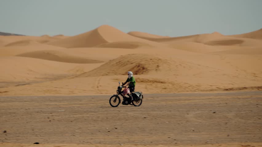 A man rides a motorcycle through the vast Sahara Desert in Morocco. He wears a headscarf for protection from the sun and sand. The journey is adventurous and scenic, beauty of the desert landscape.