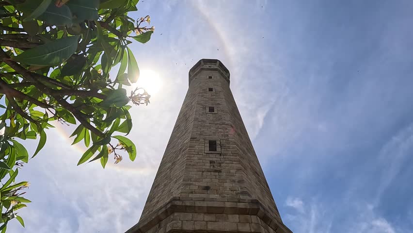 Looking up at Ke Ga Lighthouse in La Gi town, Vietnam, with a glowing sun halo overhead and scattered clouds in the sky.