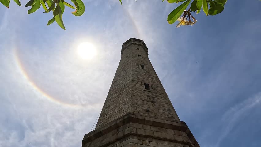 Looking up at Ke Ga Lighthouse in La Gi town, Vietnam, with a glowing sun halo overhead and scattered clouds in the sky.
