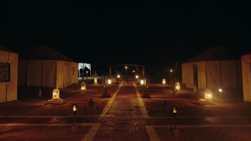 Tranquil desert night scene at a Moroccan campsite with glowing lanterns illuminating the path between traditional tents. A peaceful and inviting atmosphere of adventure and tranquility.
