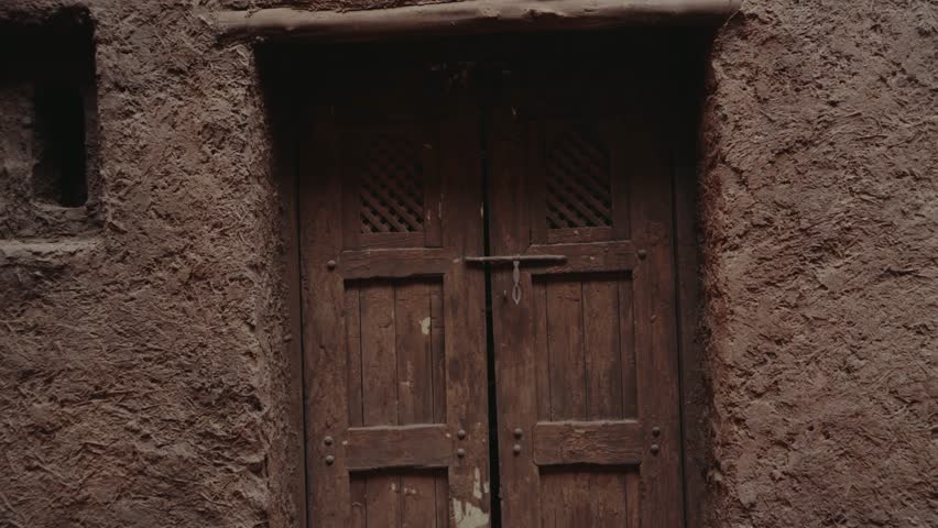 Old wooden door in a traditional Moroccan house. The rustic texture of the mud walls and the weathered wood creates a sense of history and timelessness.