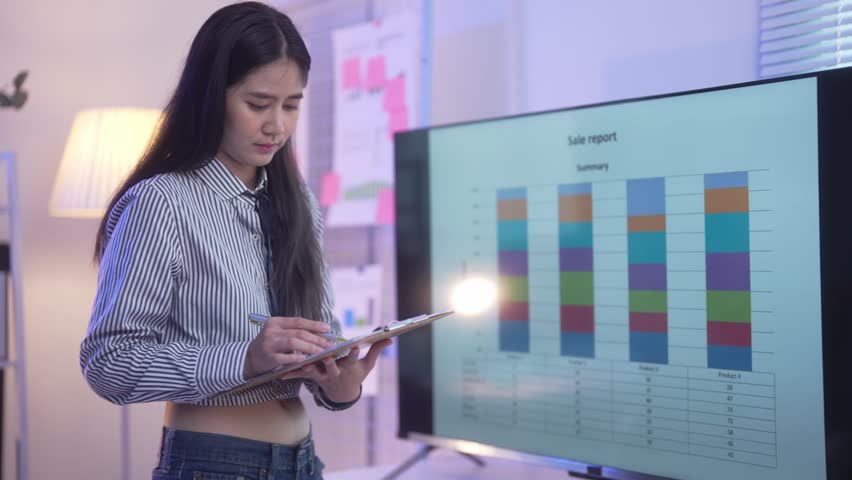 A woman is looking at a computer monitor with a slide presentation. She is writing on a clipboard