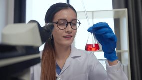 Scientist in laboratory examines red liquid in flask with microscope, showcasing dedication, focus, and curiosity in her chemistry research experiment while wearing gloves. - Powered by Shutterstock - Get 15% off with code: PIKWIZARD15