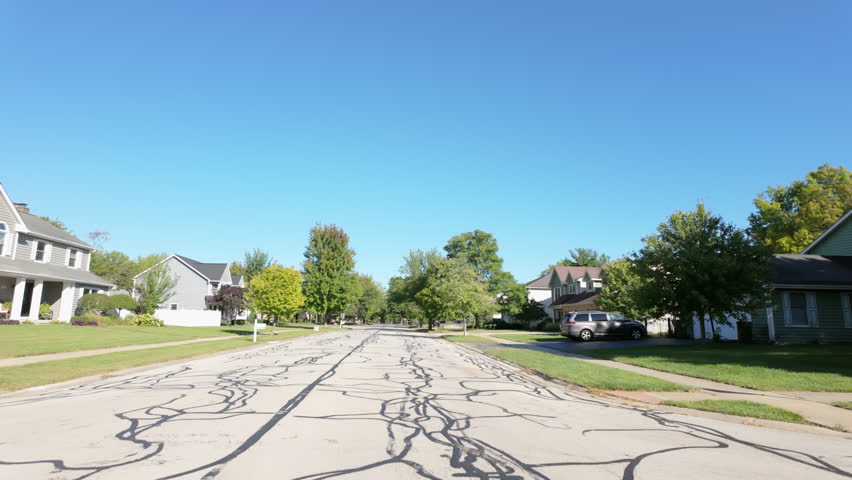Wide POV shot taken from a moving car, capturing a residential neighborhood near a park in the U.S. on a bright and sunny morning.