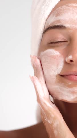 Caucasian woman with a towel on her head gently massages foaming cleanser onto her skin. Ideal concept for facial care, skincare products, beauty tutorials, and wellness advertising.