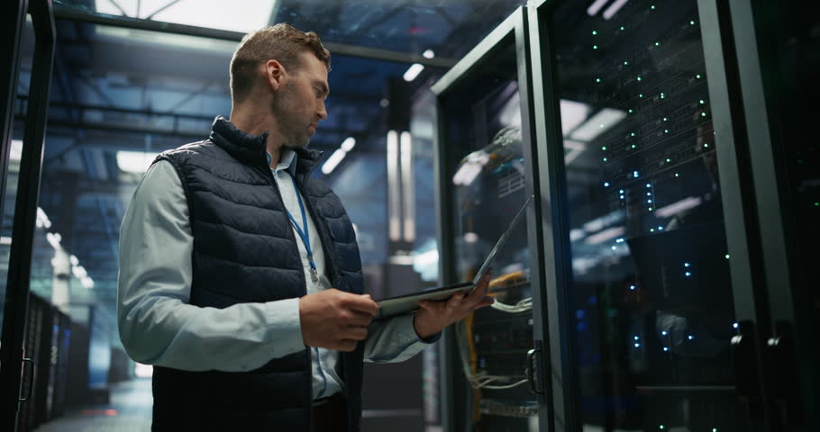 Male Data Center IT Engineer Standing in a Room with Server Racks. Cloud Computing Architect Using Laptop Computer for Servicing the System in Cyber Security and Data Protection Facility