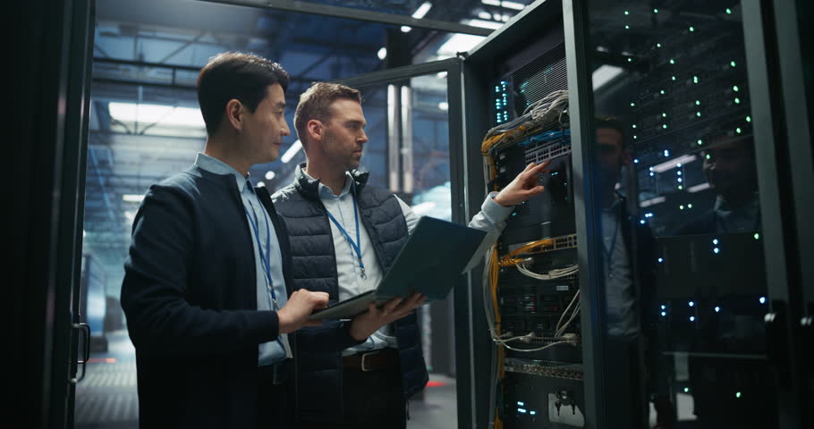 Asian IT Technician with a Laptop Computer and Caucasian Male Engineer Colleague are Talking in Data Center while Working Next to Server Racks. Running Diagnostics and Doing Maintenance Work