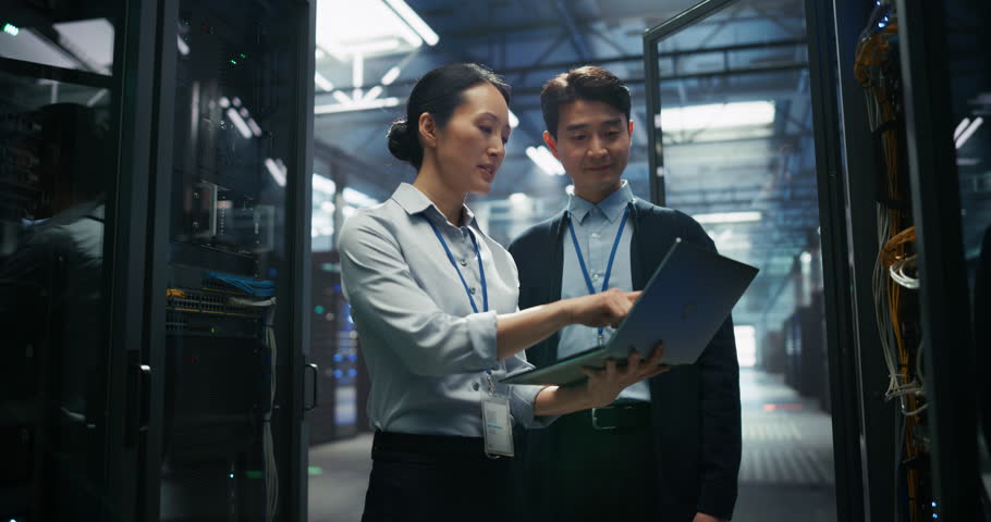 Asian Data Center Female eBusiness Entrepreneur and Male IT Specialist Having a Conversation and Using Laptop Computer. Two Information Technology Engineers Discussing Cloud Computing Server Farm