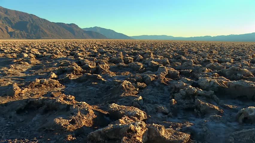 Wide view of jagged, crusty salt formations stretching across flat desert basin with distant mountains under golden light in Death Valley National Park, California, USA