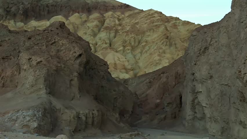 Layered eroded canyon walls with pale yellow and red rock formations framed by steep cliffs in Death Valley National Park, California, USA. Zoom out view