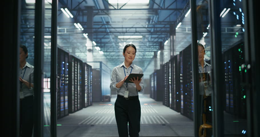 Portrait of an Asian Female IT Manager Using Tablet Computer, Walking Between Server Rack Cabinets and Supervising Data Center Operation. Male Pushing a Cart in the Background. Slow Motion Footage