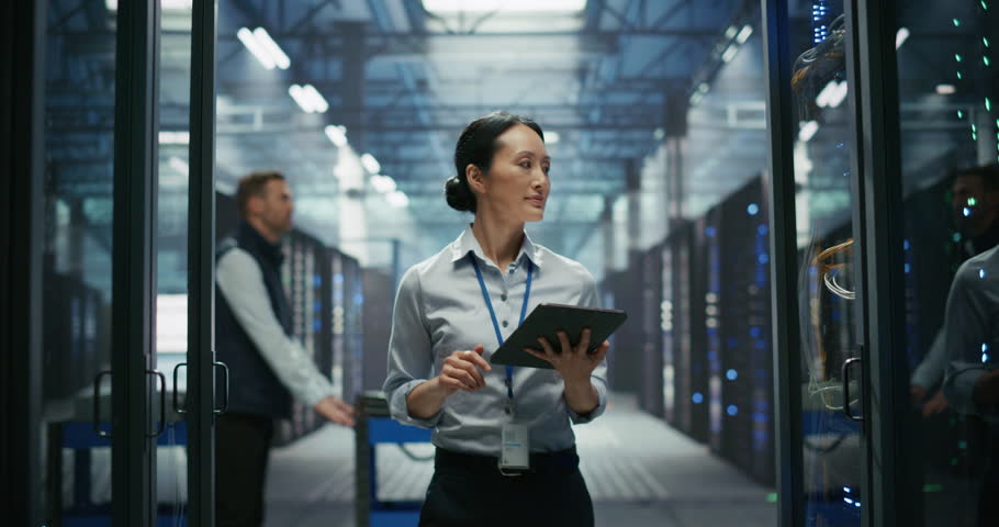 Portrait of an Asian Female IT Manager Using Tablet Computer, Walking Between Server Rack Cabinets and Supervising Data Center Operation. Male Pushing a Cart in the Background. Slow Motion Footage