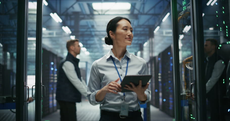 Asian Female AI Engineer Using Tablet Computer while Walking Between Server Racks and Inspecting Hardware and Stable Connection. Colleague Pushing a Cart in the Background. Slow Motion Footage