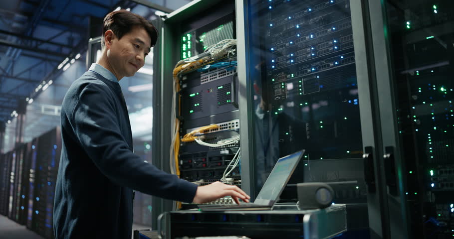 Data Center: Asian Programmer Using Laptop Computer, Focused while Inputting Data. Cloud Computing Server Farm System Administrator Working on IT Maintenance for Saas Business Company