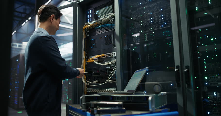Asian Server Technician Installing Additional Hardware into a Server Cabinet in a Data Center. Specialist Running Diagnostics on a Laptop Computer, Optimizing Storage Hard Drives in a Modern Facility