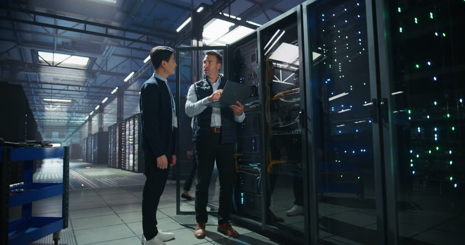 Asian IT Technician and Caucasian Male Engineer with a Laptop Computer are Talking in Data Center while Working Next to Server Racks. Running Diagnostics and Doing System Maintenance Work