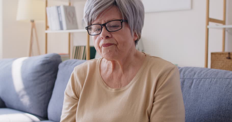 Tired elderly woman sitting on sofa and rubbing eyes while holding glasses in hand. Senior lady experiencing vision fatigue, strain or discomfort after reading or spending time in front of screen.