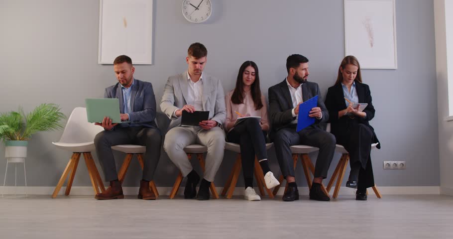 Five young business professionals sitting on chairs in row with laptop and documents. Focused adult male and female job candidates preparing and waiting for interview or business meeting in office.