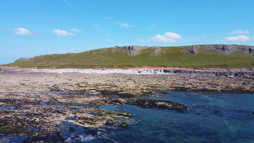 Pushing Aerial Shot Towards Welsh Coastline in The Gower Peninsula with Clear Water Rock Pools on Rocky Beach and Coastal Paths with Steep Cliffs and Blue Sky. Ocean Nature Footage.