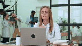 Stressed female healthcare worker typing on laptop, medical team collaborating around her in crowded hospital setting, showing professional workplace tension - Powered by Shutterstock - Get 15% off with code: PIKWIZARD15