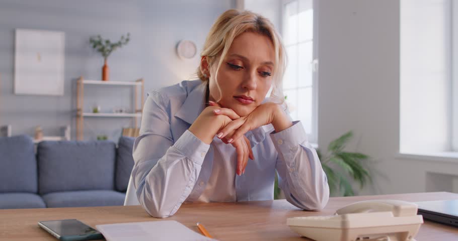 Portrait of unhappy young sad woman in casual clothes sitting at the desk at home waiting for important phone call from someone. Desperate female person looking on telephone indoors. 4k video.