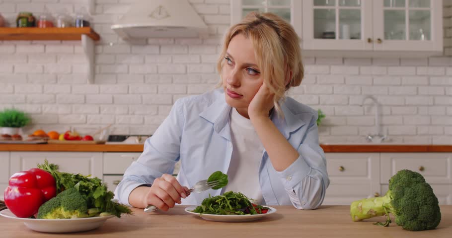 Unhappy young woman eating unwillingly salad, keeping to diet, consuming fresh green vegetables, sitting on table against kitchen interior. People, nutrition and lifestyle concept. 