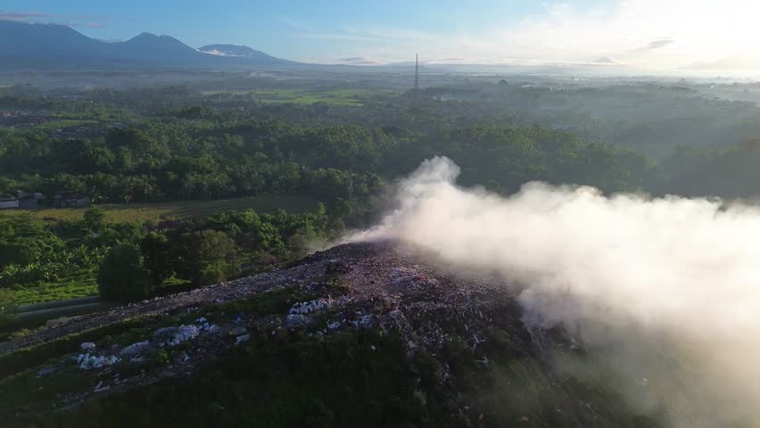 Aerial view of open trash burning at a rural landfill. Smoke rises into the air with scenic mountains in the background, highlighting the contrast between pollution and nature