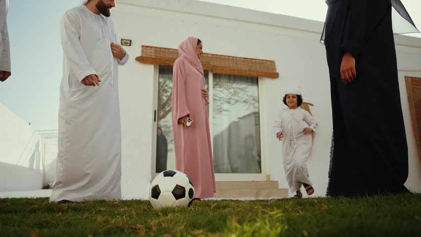 Arab family, dressed in traditional clothing, enjoys a sunny day playing soccer together in their lush green garden, fostering family bonds and creating cherished memories